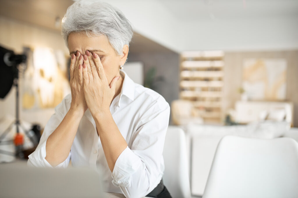Senior sad woman with gray hair working on laptop, rubbing eyes or hiding tears, full of restless thoughts, having problem. Frustrated mature female being stressed because of financial trouble